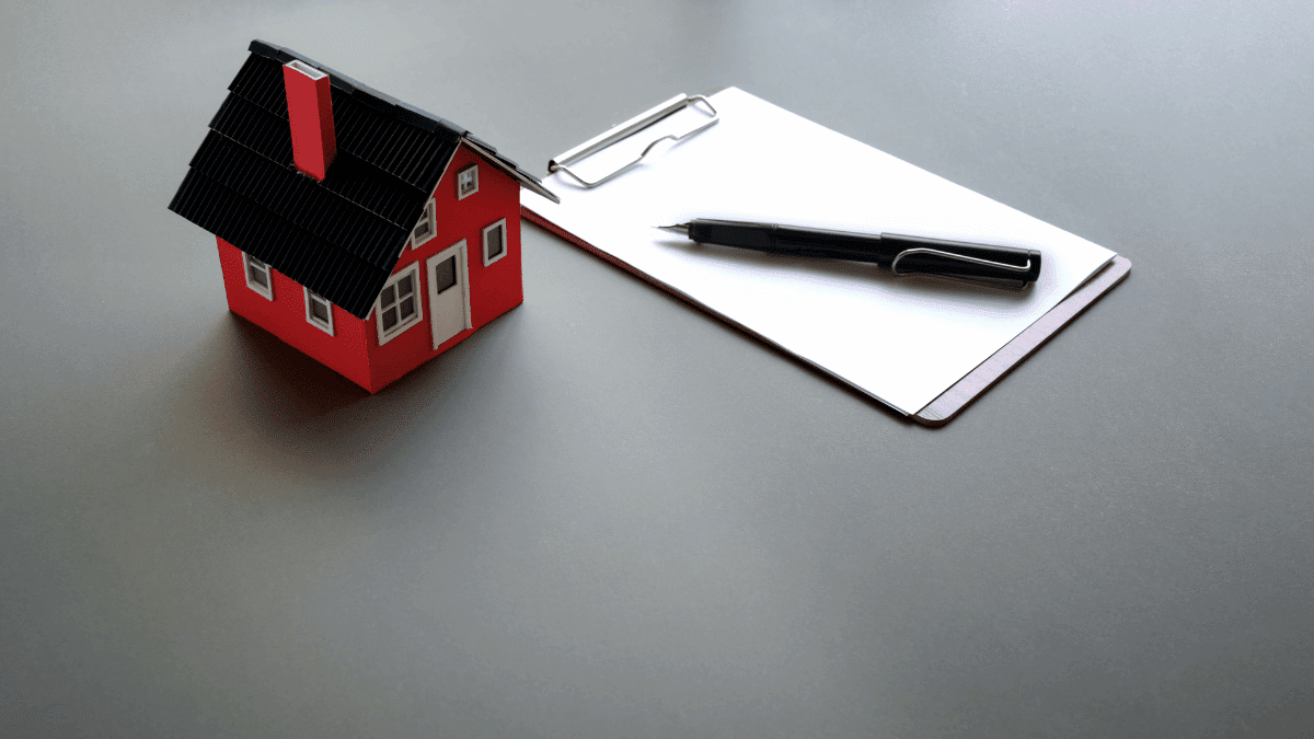 Imagem de uma miniatura de casa e um contrato ao lado com uma caneta em cima. Ambos estão em cima de uma mesa.