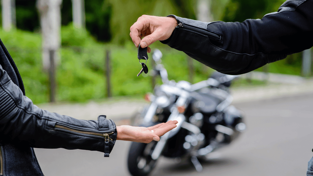 imagem de um homem dando a chave de uma moto para uma mulher (ambos apenas mostrando o braço e mãos) em uma rodovia com uma moto ao fundo.