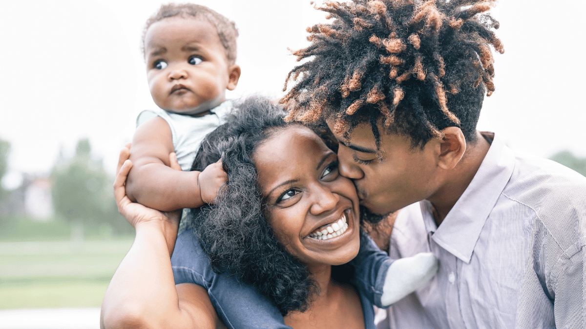 Imagem de uma família negra composta por um homem, uma mulher e um bebê que está sobre os ombros da mulher enquanto o homem beija o rosto da mulher.