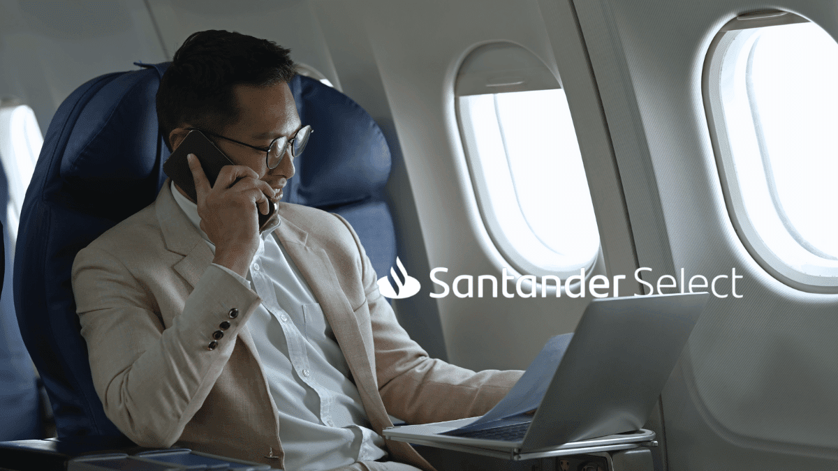 Imagem de um homem com traços asiáticos em um assento de avião. Ele está usando paletó bege, sorrindo falando ao telefone com um laptop no colo. Ao lado dele está o logo do Santander Select.